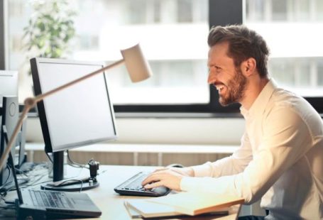 Dual Monitors - Man in White Dress Shirt Sitting on Black Rolling Chair While Facing Black Computer Set and Smiling