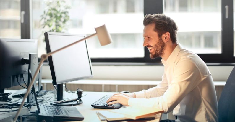 Dual Monitors - Man in White Dress Shirt Sitting on Black Rolling Chair While Facing Black Computer Set and Smiling