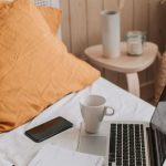 Portable Computer - From above of open netbook with black screen and cup of coffee with smartphone and notebook placed on crumpled bed sheet in bedroom with big vase on floor