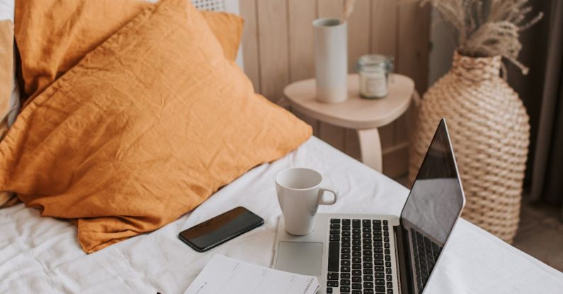 Portable Computer - From above of open netbook with black screen and cup of coffee with smartphone and notebook placed on crumpled bed sheet in bedroom with big vase on floor