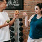 Connectivity Options - Side view of Asian personal instructor with apple and doughnut offering healthy food to plus size woman while standing in gym