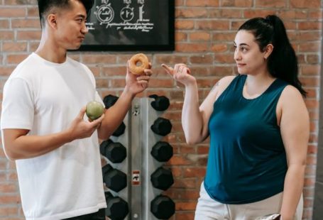 Connectivity Options - Side view of Asian personal instructor with apple and doughnut offering healthy food to plus size woman while standing in gym