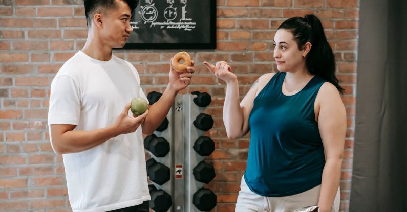 Connectivity Options - Side view of Asian personal instructor with apple and doughnut offering healthy food to plus size woman while standing in gym